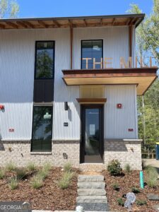 Modern two-story building with corrugated metal siding, black-framed glass doors/windows, and a wooden sign spelling 'THE MILL' above the entrance; manicured front landscaping with mulch and grasses.