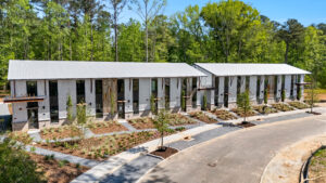 Modern townhouses line a curved street with white metal siding, large windows, and landscaped front yards against a wooded backdrop.