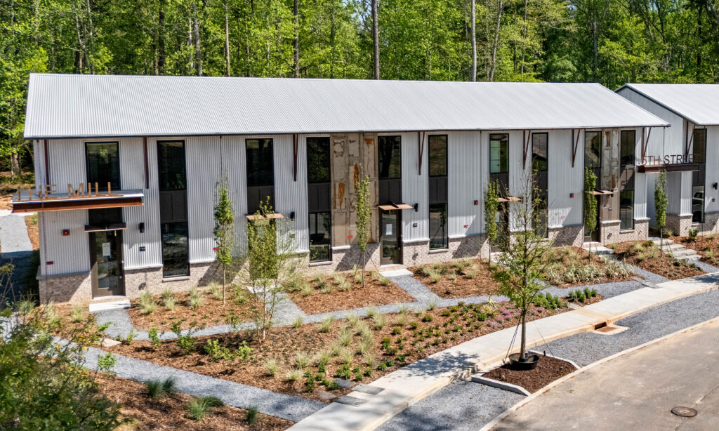 Row of modern townhouses with gray metal siding, stone bases, large windows, and landscaped front yards along a sidewalk.