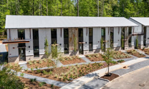 Row of modern townhouses with gray metal siding, stone bases, large windows, and landscaped front yards along a sidewalk.